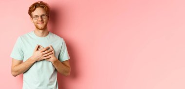 Kızıl saçlı çekici bir genç adam, kalbini okşuyor ve gülümsüyor. Minnettar, teşekkür ediyor, pembe arka planda duruyor..