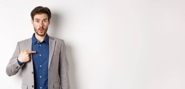 Kim ben mi? Takım elbiseli şaşırmış bir adam kendini inançsızlıkla işaret ediyor, kameraya bakıyor, beyaz arka planda duruyor..