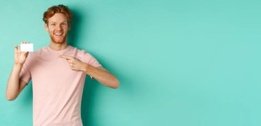Sakallı ve kızıl saçlı çekici yetişkin adam plastik kredi kartını işaret ediyor, kameraya gülümsüyor, turkuaz arka planda duruyor..