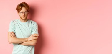 Gözlüklü, kızıl saçlı, kollarını göğsüne dayamış, kendini savunurken sana somurtan, pembe arka planda duran kızgın bir adam..