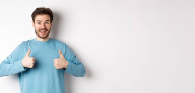 Heyecanlı gülümseyen adam mükemmel ürünü övmek için başparmağını kaldırıyor, kameraya şaşkın ve mutlu bakıyor, iyi bir anlaşma öneriyor, beyaz arka planda duruyor..