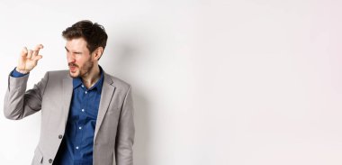 İş adamı küçük bir şey görmeye çalışıyor, parmaklarıyla küçük bir şey gösteriyor, beyaz arka planda takım elbise içinde duruyor..