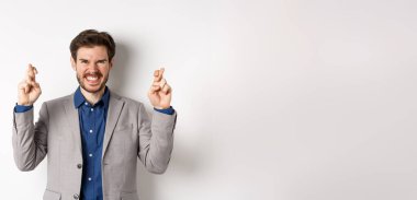 Motive olmuş ve heyecanlı iş adamı pozitif gülümsüyor, umut dolu görünüyor ve şans diliyor, dilek tutuyor, duyuruyu bekliyor, beyaz arka plan.