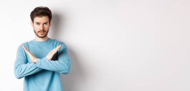 Sakallı genç adam ciddi görünüyor, bir şeyi durdurmak için çapraz jest yapıyor, eylemi yasaklıyor, beyaz arka plana karşı duruyor..