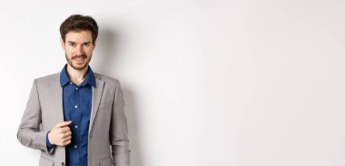 Takım elbiseli kendine güvenen işadamı kameraya gülümsüyor, beyaz arka plana karşı kendinden emin duruyor..