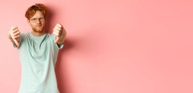 Gözlüklü, dağınık kırmızı saçlı hayal kırıklığına uğramış genç adam, baş parmaklarını aşağı gösteriyor ve tatminsiz yüzünü ekşitiyor, pembe arka planda durmaktan hoşlanmadığını ifade ediyor..