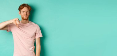 Hayal kırıklığına uğramış kızıl saçlı adam, baş parmağını kaldırmış, kaşlarını çatmış, şüpheci, hoşnutsuz ve hoşnutsuz görünüyor, turkuaz arka planda duruyor..