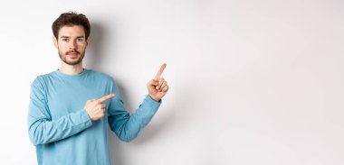 Beyaz arka planda sıradan kıyafetler giymiş, logoyu işaret eden ve kameraya bakan ciddi bir beyaz adamın portresi..