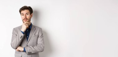 Takım elbiseli düşünceli bir işadamı, sakala dokunuyor, düşünüyor ve kameraya bakıyor, düşünceli bir yüzle karar veriyor, beyaz arka planda duruyor..