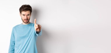 Sakallı, parmağını sallayarak onaylamadığı, bir şeyi yasakladığı ya da yasakladığı beyaz arka planda duran ve hayır diyen ciddi bir gencin görüntüsü..