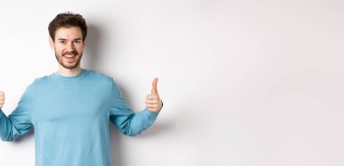 Neşeli genç adam başparmağını kaldırıyor ve gülümsüyor, iyi ürün öneriyor, seçimi övüyor, beyaz arka planda duruyor..