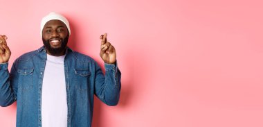 İyimser Afro-Amerikalı bir adam dilek diliyor, parmaklarını çapraz tutuyor ve gülümsüyor, pembe arka planda duruyor..