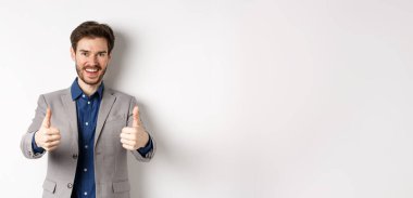 Takım elbiseli neşeli ve pozitif genç adam iyi bir şey gibi başparmaklarını kaldırıyor ve gülümsüyor, iyi bir iş çıkarıyor, beyaz arka planda mutlu duruyor..