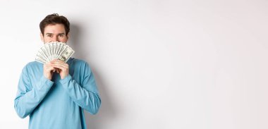 Neşeli alışverişçi alışveriş için para tutuyor, dolarlarla ayakta duruyor ve gülümsüyor, günlük kıyafetlerle beyaz arka planda duruyor..