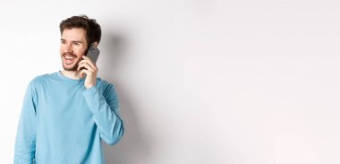 Teknoloji konsepti. Neşeli adam telefonla konuşmaktan hoşlanıyor, akıllı telefondan konuşuyor ve gülümsüyor, beyaz arka planda sıradan bir tişörtle dikiliyor..