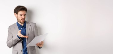 Kızgın işveren kötü belgeye bakıyor, anlaşmadan şikayetçi, kağıttan bıkmış, beyaz arka planda takım elbise içinde duruyor..