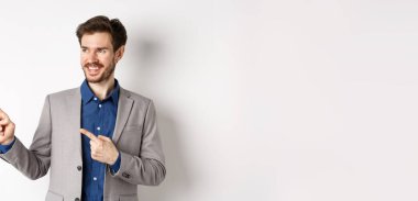 Gri takım elbiseli başarılı bir işadamı solu işaret ediyor ve pankarta bakıyor, kendinden emin gülümsüyor, reklamını yapıyor, beyaz arka planda duruyor..