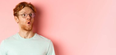 Kızıl saçlı, sakallı, kaşlarını kaldıran, şaşırmış ve pembe arka planda duran sağ üst logoya bakan meraklı genç adamın yüzü..