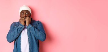 Korkmuş siyah adam korkudan titriyor, tırnaklarını yiyor ve kameraya bakıyor, pembe arka planda dikiliyor..