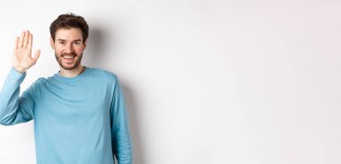 Sakallı neşeli genç adam merhaba diyor, dostça bakıyor ve el sallıyor, beyaz arka planda duruyor..