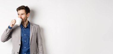 Takım elbiseli yakışıklı bir işadamı ofis kupasından kahve ya da çay içiyor, logoya bakıyor, beyaz arka planda duruyor..