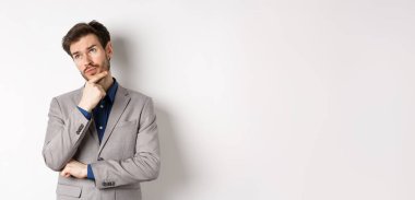 Gri takım elbiseli düşünceli bir işadamı sol üst köşedeki logoya bakar, düşünür ya da karar verir, plan yapar, beyaz arka plana karşı durur..