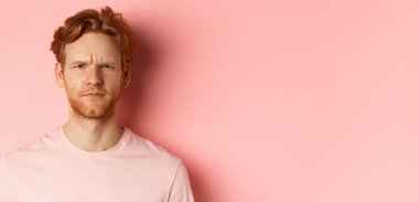 Şüpheli, kaşlarını çatmış, kameraya şüpheyle bakan, onaylamadığını ifade eden, pembe arka planda duran kızıl saçlı adamı kapat..