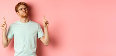 Gözlüklü, kaşlarını çatan, hayal kırıklığına uğramış, pembe arka planda duran bir şeyi işaret eden şüpheli kızıl saçlı adam..