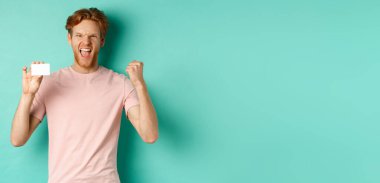Neşeli genç adam zafer kazanıyor, başarıyı kutlamak için yumruk tokuşturuyor, plastik kredi kartı gösteriyor, bankadan ödül kazanıyor, turkuaz arka plan.