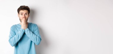 Şok olmuş ve endişeli genç adam soluk soluğa ve ağzı kapalı, kameraya panikle bak, beyaz arka plana karşı endişeli dur.