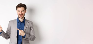 Beyaz takım elbiseli yakışıklı beyaz adam sol pankartı işaret ediyor ve gülümsüyor, reklam yapıyor, beyaz arka plana karşı duruyor..