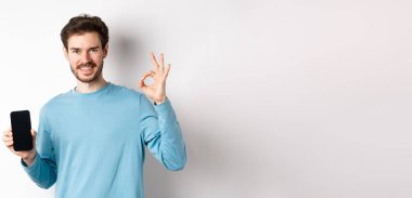 E-ticaret ve alışveriş konsepti. Yakışıklı sakallı adam boş cep telefonu ekranı ve tamam işareti gösteriyor, iyi uygulamayı övüyor, beyaz arka planda duruyor..