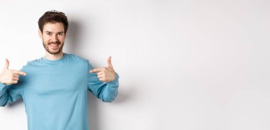 Yakışıklı sakallı adam, parmaklarını merkeze doğrultuyor, kendi reklamını yapıyor ve kendinden emin görünüyor, beyaz arka planda duruyor, kameraya gülümsüyor..