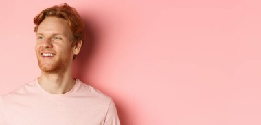 Sakallı, kızıl saçlı, soluna bakıp gülümseyen pembe arka planda kendine güvenen yakışıklı bir genç..