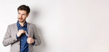 Kendine güveni tam ve başarılı bir iş adamı gibi görünen ve beyaz arka planda duran plastik kredi kartını takım elbisenin cebine koyan..