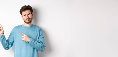 Şüpheci ve etkilenmemiş genç adam, yüzünü ekşitiyor, kötü terfiye bakıyor, beyaz arka planda tatminsiz duruyor..