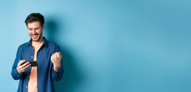 Cep telefonuyla video oyununda kazanan mutlu genç adam, akıllı telefon ekranına bakıyor ve sevinçle evet diyor, mavi arka planda duruyor..