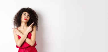 Kırmızı elbiseli, çapraz parmaklı, yana bakan, dalgın dalgın bakan, seçim yapan, beyaz arka planda duran kararsız budala kadın..