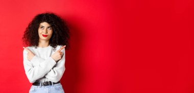 Düşünceli genç bir kadın seçim yapıyor, parmaklarını yana çeviriyor ve kendine güvenen yüzüyle sol üst köşeye bakıyor, kırmızı arka planda duruyor..