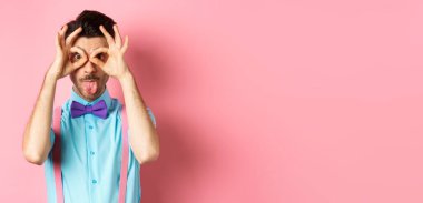 Papyon ve pantolon askısı takmış, el dürbününden bakıp kameraya dilini gösteren, pembe arka planda neşeli duran komik ve aptal genç bir adam..