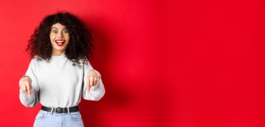 Heyecanlı kadın parmaklarını aşağıya doğru gösteriyor ve şaşkın bir şekilde gülümsüyor, şuraya bakın, kırmızı boşlukta logo gösteriyor..