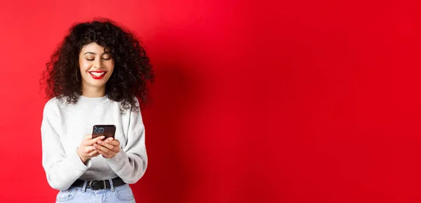 Happy positive girl chatting on phone, reading message and smiling, using social media app, standing on red background.
