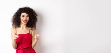 Kırmızı elbiseli güzel kadın seni tebrik ediyor, kamerayı işaret ediyor ve gülümsüyor, beyaz arka planda duruyor..