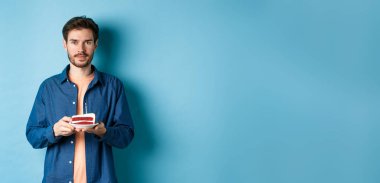 Doğum gününü kutlayan gülümseyen genç bir adam. Elinde mumla doğum günü pastası tutarken mavi arka planda dikilip kameraya bakarken..