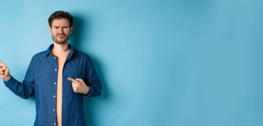 Hüsrana uğramış ve şüpheci adam somurtuyor, dudaklarını büzüyor ve parmaklarını boş yere doğrultuyor, kötü şeyden yakınıyor, mavi arka planda üzgün duruyor..