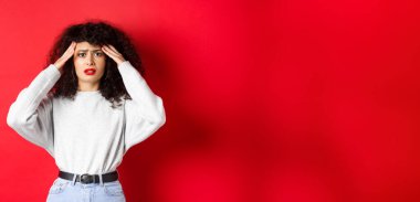 Endişeli ve endişeli genç bir kadın kafasına dokunuyor ve yüzünü buruşturuyor, baş ağrısı çekiyor, kırmızı arka planda migrenle ayakta duruyor..