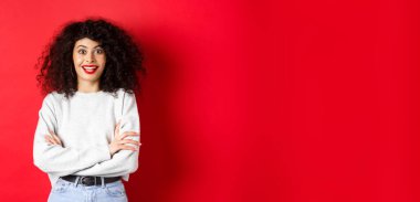 Kıvırcık saçlı neşeli genç bir kadın, kaşlarını kaldırıyor ve şaşırmış görünüyor, ilginç haberler duyuyor, kırmızı arka plan.