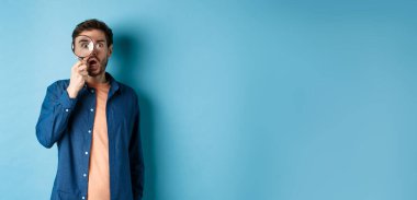 Etkilenmiş bir adam büyüteçle huşu içinde bakar, çenesini indirir ve kameraya bakar, mavi arka planda durur..