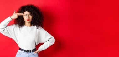 Kıvırcık saçlı, kafasında silah işareti olan, kafası patlayan, kırmızı arka planda duran, rahatsız bir beyaz kadın..