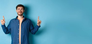 Şüpheci genç adam sırıtıyor ve şüpheli bakıyor, üzgün parmaklarını gösteriyor, mavi arka planda duruyor. Boşluğu kopyala